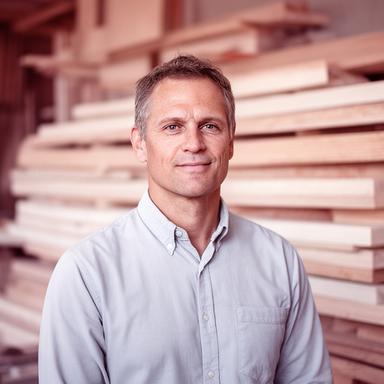 Simon Baumann vor gestapelten Holzbohlen in einer Werkhalle mit natürlichem Licht